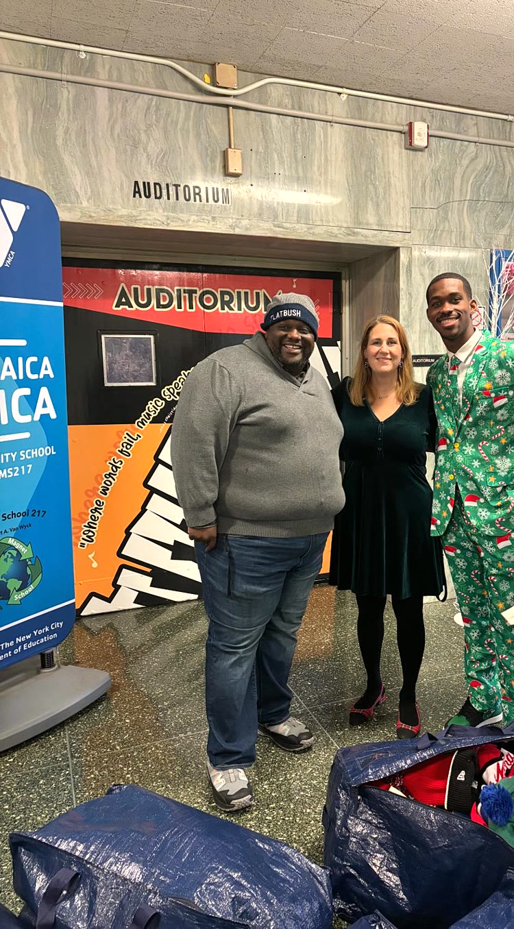 Three adults smile in a school lobby beside large bags of donated winter hats, in front of the auditorium doors at JHS 217.
